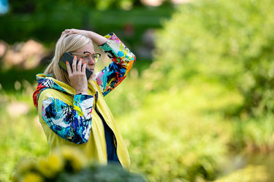 Brightly dressed blonde senior woman talking on smartphone in garden, online communication concept