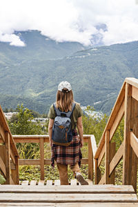 Rear view of young blonde woman looking at view of mountains standing on wooden viewpoint, hiking 