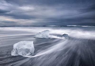 Scenic view of icebergs on beach with rough sea against cloudy sky
