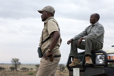 Male friends by off-road vehicle against cloudy sky at serengeti national park