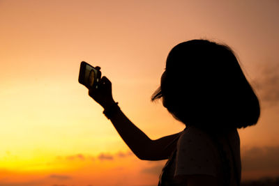 Side view of young woman taking selfie with smart phone against sky during sunset