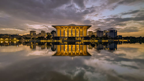 Illuminated building by lake against sky during sunset