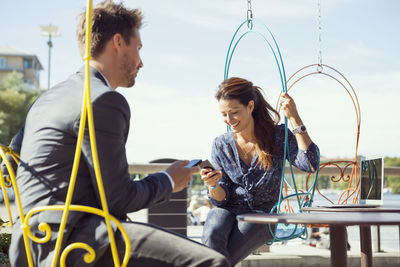 Happy businesswoman using mobile phone on swing while sitting with colleague at sidewalk cafe