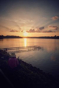 Rear view of man sitting by lake against sky during sunset