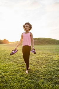 Portrait of happy girl holding sports shoe on grass against sky during sunset