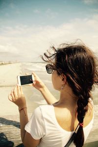 Rear view of woman photographing with mobile phone against beach