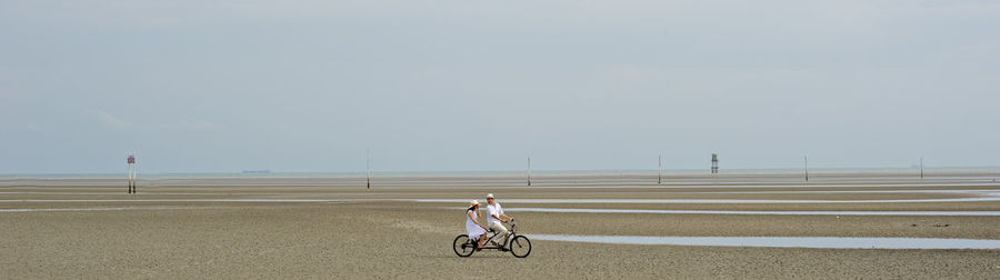 Panoramic view of man with woman riding bicycle at sandy beach against sky