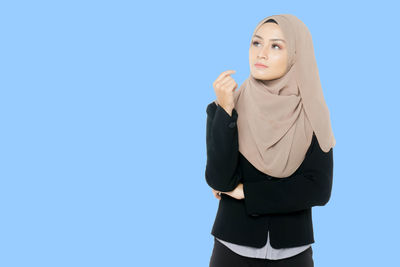 Thoughtful young woman wearing hijab while standing against blue background