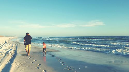 Father and son walking on sea shore at beach against blue sky