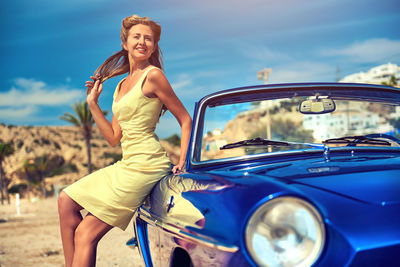 Portrait of smiling young woman sitting on car against blue sky