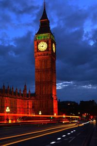 Light trail on street by illuminated big ben against cloudy sky