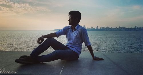 Man sitting by sea against sky during sunset