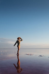 A young caucasian woman in a sweater, hat and short shorts walks barefoot on a pink salt lake.