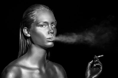 Close-up portrait of young woman with body paint smoking against black background