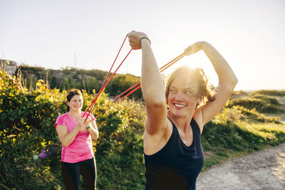 Smiling woman holding resistance band while exercising with woman at beach