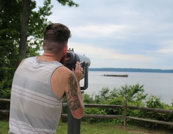 Rear view of man holding cigarette while standing by coin-operated binoculars against lake and sky