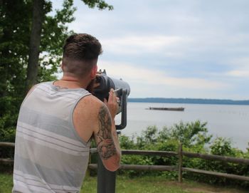 Rear view of man holding cigarette while standing by coin-operated binoculars against lake and sky