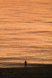 Silhouette man fishing while standing at shore during sunrise