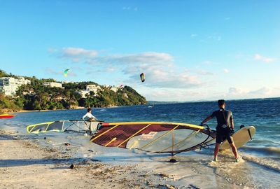 People with windsurfing board on shore at beach against sky