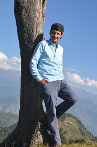 A happy young guy looking at camera while posing with leaning against tree in hilly area 