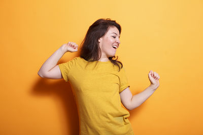 Portrait of young woman with arms raised against yellow background