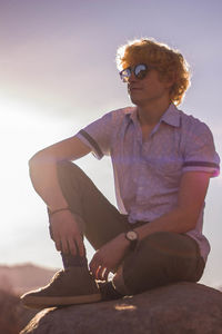 Man in sunglasses sitting on rock against sky during sunset