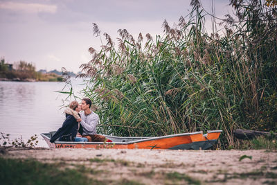 Side view of young woman sitting by lake against sky