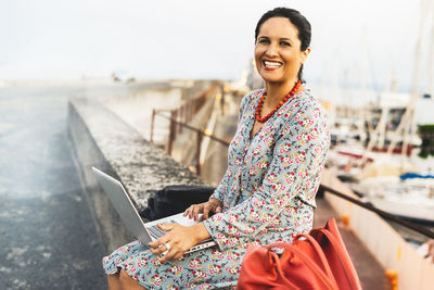 Portrait of cheerful woman using laptop while sitting at harbor