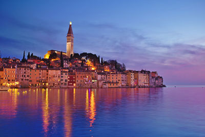 Illuminated buildings by sea against sky at dusk