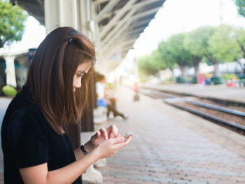 Side view of young woman using phone while sitting on railroad station