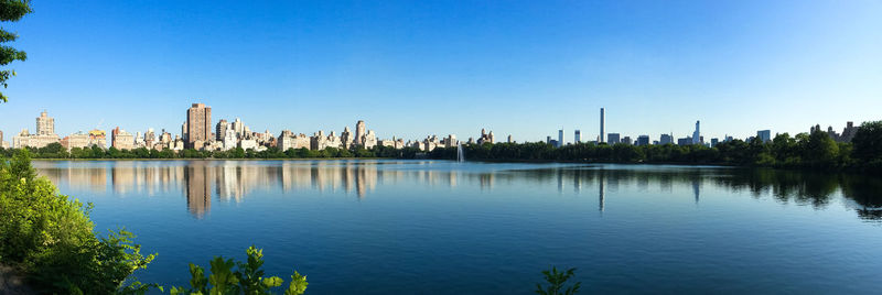 Panoramic view of buildings in city against clear blue sky reflecting on pond at central park