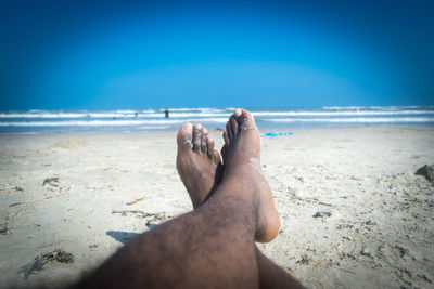 Low section of man relaxing on sand at beach against blue sky