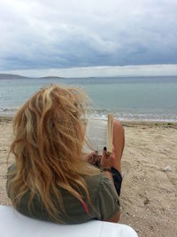 Woman with blond hair reading book at beach against cloudy sky