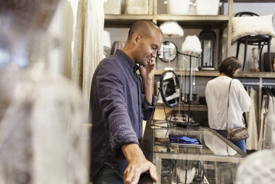 Smiling owner using phone while female customer looking at clothes in store