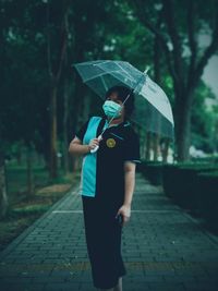 Woman wearing surgical mask while holding umbrella on footpath during rainy season