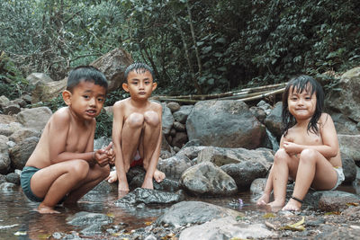 Full length cute kids sitting by rocks in forest