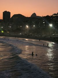 Silhouette people on beach by illuminated city against sky at sunset