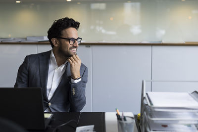 Smiling male real estate agent looking away while sitting with hand on chin in office