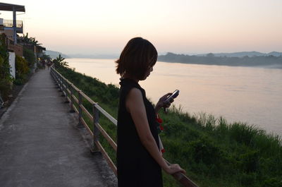 Side view of woman using phone on footpath by river