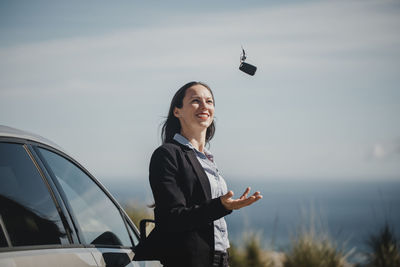 Smiling entrepreneur playing with car key while standing against sky