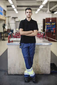 Full length portrait of confident male auto mechanic student standing arms crossed in workshop