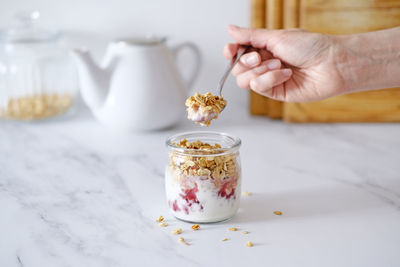 Female hand holds a spoon of granola with yogurt and red berries in a glass jar