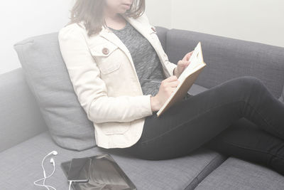 Midsection of woman writing in diary while sitting by smart phone and digital tablet on sofa at home
