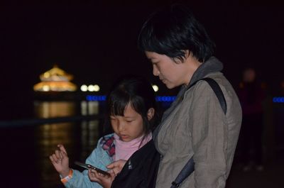 Mother and daughter using mobile phone while standing by lake at night