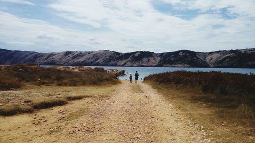 Rear view of father and child walking on dirt road towards lake against cloudy sky