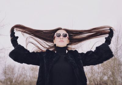 Low angle view of young woman wearing heart shape sunglasses while standing against clear sky during winter