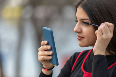 Outdoors portrait of young beautiful teen using smartphone as mirror while do her makeup