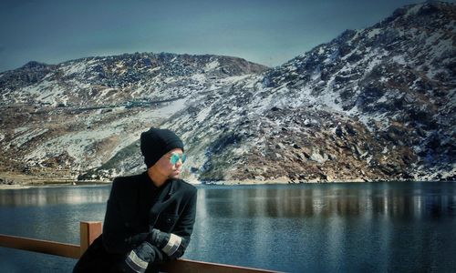 Young man leaning on railing by lake against mountain range