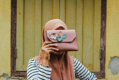 Woman holding purse over face against wall