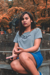 Portrait of young woman using phone while sitting on steps against autumn trees